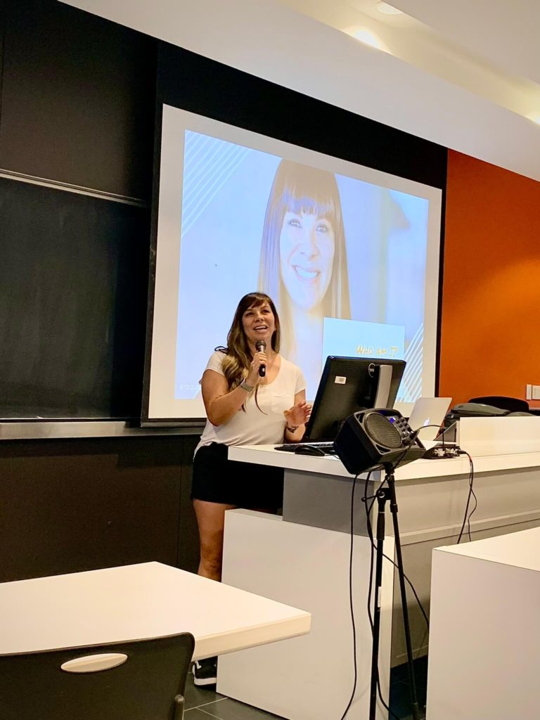 raquel standing behind desk/podium, in front of a big screen, giving a talk